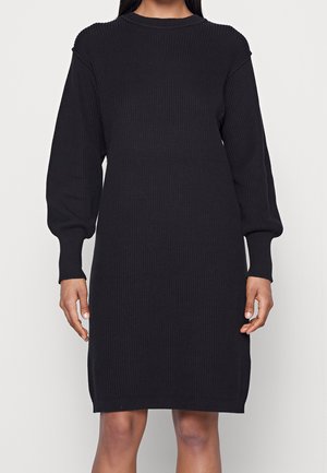 Vrouw die een zwarte, geribbelde gebreide jurk met lange mouwen en knielengte draagt tegen een effen achtergrond.