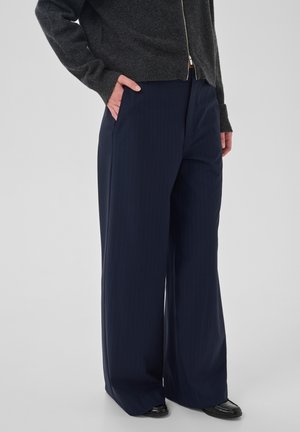 Persona con pantalones oscuros de pierna ancha, suéter oscuro con cremallera y zapatos negros, de pie con una mano en el bolsillo frente a un fondo liso.