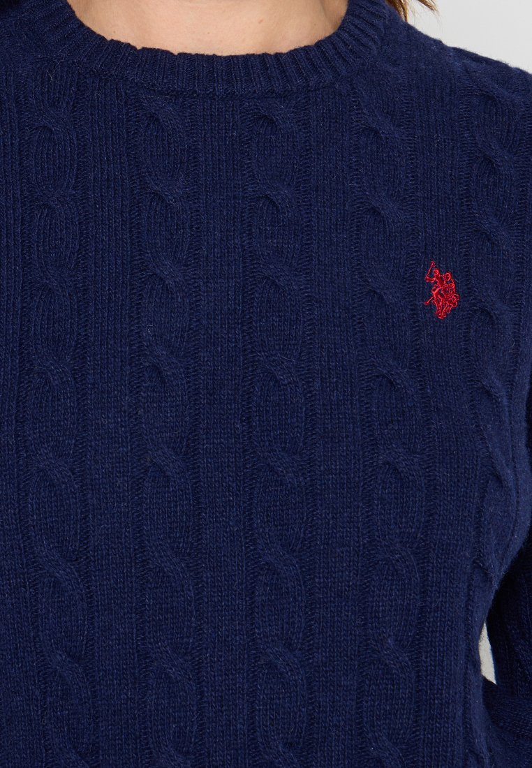 Navyblauer Kabelstrickpullover mit rundem Ausschnitt und langen Ärmeln. Kleines rotes gesticktes Wappen auf der Brust. Weicher Wollstoff.