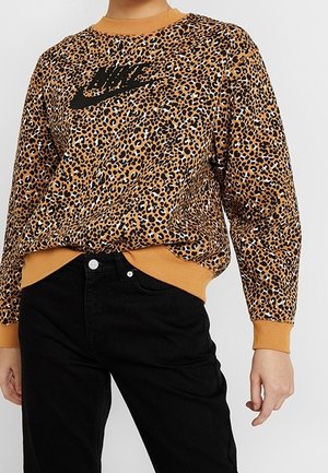Sweat-shirt Nike à manches longues imprimé léopard avec poignets et ourlet côtelés couleur moutarde, porté avec un pantalon noir taille haute.