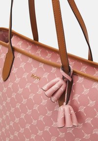 Vaaleanpunainen tekstuurinen olkalaukku, jossa on toistuva kuvio, ruskeat nahkahihnat ja kaksi yhteensopivaa tupsua. Sisältää hillityn kultaisen logon yksityiskohdan.