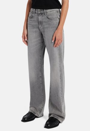 Person iført grå jeans med høj talje og løs pasform samt sorte sko med firkantet tå, stående mod en ensfarvet lys baggrund.