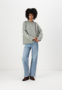 Hellgrüner oversized Pullover mit geripptem Saum, kombiniert mit hochtaillierten hellblauen geraden Jeans. Braune Absatzstiefeletten runden das Outfit ab.