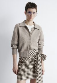 Hellbrauner Wollcardigan mit Kragen, Knopfleiste und gerippten Bündchen; kombiniert mit einem schwarz-weißen karierten Wickelrock.