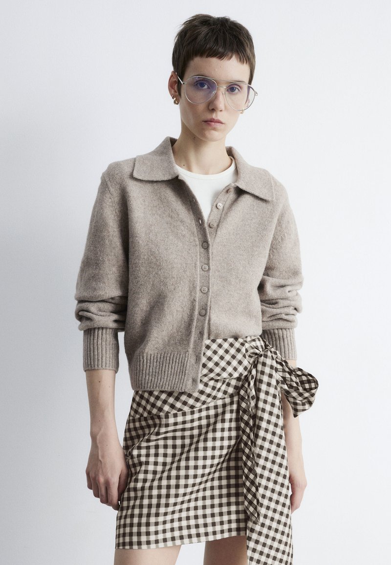 Hellbrauner Wollcardigan mit Kragen, Knopfleiste und gerippten Bündchen; kombiniert mit einem schwarz-weißen karierten Wickelrock.