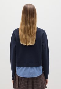 Navy-blauer gestrickter Pullover mit Rundhalsausschnitt, der über einem hellblau gestreiften Hemd getragen wird, kombiniert mit einem braunen Karorock mit Falten.