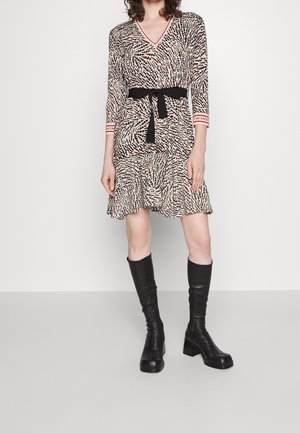 Vrouw draagt een beige jurk met rood-zwarte vlekken, een zwarte taille riem en zwarte kniehoge laarzen, staand tegen een effen achtergrond.