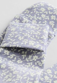 Tissu violet clair avec un motif floral blanc. Texture douce, design tricot, mettant en avant de petites feuilles et fleurs. Bord plié visible.