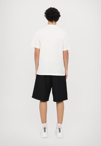 Tricou alb din bumbac, cu croială lejeră, asociat cu pantaloni scurți negri până la genunchi și adidași albi cu dungi negre, văzut din spate.