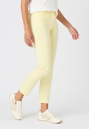 Personne portant un pantalon ajusté jaune clair et des baskets blanches avec des accents léopard dorés, debout devant un fond uni.