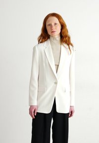 Blazer bianco su misura con un solo bottone, revers a notched e due tasche frontali, indossato sopra un dolcevita beige. Pantaloni neri a gamba larga.