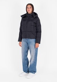 Giubbotto piumino nero con cappuccio, chiusura con zip e design imbottito. Abbinato a jeans a gamba larga di colore azzurro chiaro e sneakers bianche.