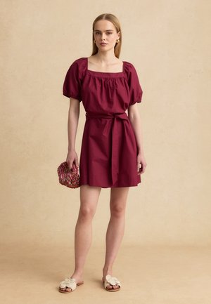 Donna che indossa un vestito bordeaux con maniche a sbuffo e cintura in vita, che tiene una pochette rosa intrecciata e indossa sandali crema a fiori senza lacci.