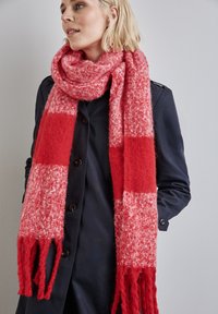 Sciarpa a quadri rossa e rosa con estremità sfrangiate, realizzata in un materiale morbido e testurizzato, indossata con un cappotto scuro.