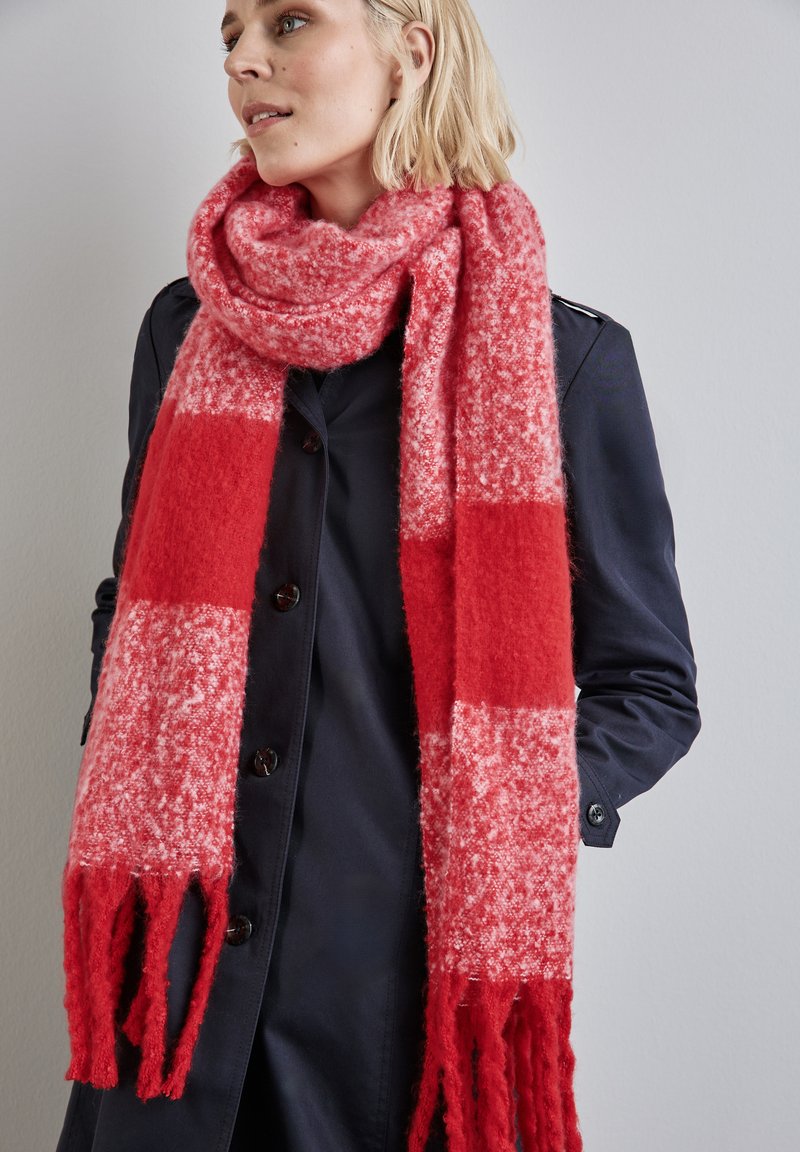 Sciarpa a quadri rossa e rosa con estremità sfrangiate, realizzata in un materiale morbido e testurizzato, indossata con un cappotto scuro.