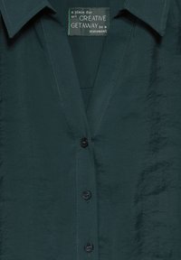 Camicia texture verde scuro con scollatura a V, colletto a punta e sei bottoni neri sul davanti. L'etichetta legge "Creative Getaway."
