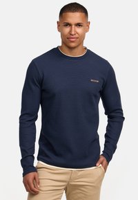 Mørkeblå langærmet sweater i ribbestrik med rund halsudskæring og et lille brunt label på brystet. Båret over lyse bukser.