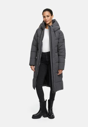 Lange grijze pufferjas met een capuchon, doorgestikte ontwerp, voorkant met ritssluiting en zijzakken, gedragen over zwarte jeans en zwarte laarzen.