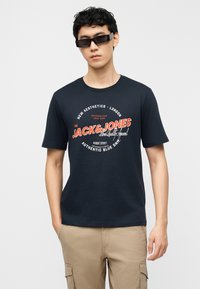 Junger Mann mit schwarzen Sonnenbrillen, marineblauem Jack & Jones Logo-T-Shirt und beigen Cargohosen, stehend vor weißem Hintergrund.