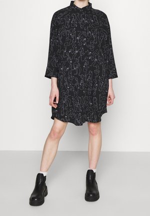Zwarte jurk met lange mouwen en abstract witte lijntekening, gedragen met witte sokken en zwarte plateau enkellaarsjes op een lichtgrijze vloer.