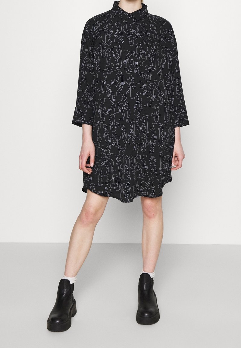 Robe noire à manches longues avec un imprimé abstrait en lignes blanches, portée avec des chaussettes blanches et des bottines à plateforme noires sur un sol gris clair.