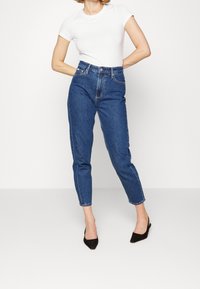 Tummansiniset korkeavyötäröiset farkut, joissa on kapeaksi kapeneva lahje, perinteinen viiden taskun malli, kontrastitikkaukset ja napillinen etumallinen kiinnitys.