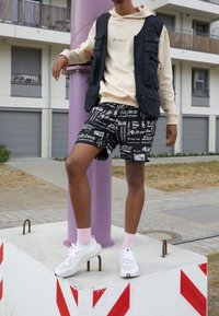 Black vest with multiple pockets, beige hoodie, black patterned shorts, white sneakers with pink laces, standing on a concrete block.
