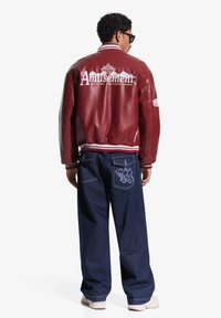 Bărbat purtând o geacă de piele roșie cu textul „Amusement” și un grafic pe spate, blugi largi de culoare albastru închis cu o emblema în formă de stea, pantofi sport albi.