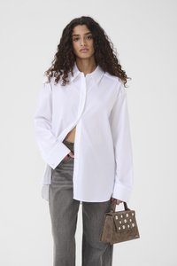 Λευκό υπερμεγέθες πουκάμισο με κουμπιά και μακριά μανίκια, συνδυασμένο με γκρι παντελόνι. Μια μικρή καφέ τσάντα από σουέντ διαθέτει λευκές κυκλικές λεπτομέρειες.