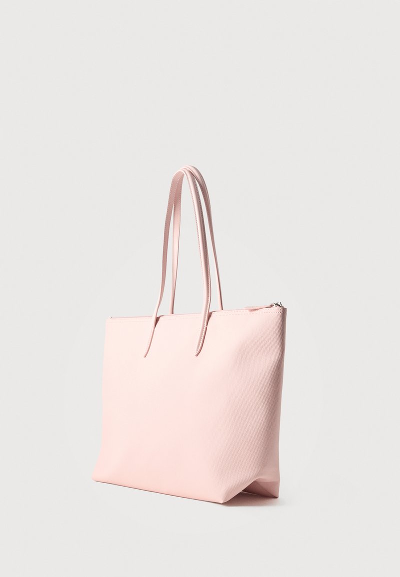 Borsa tote rosa morbida e con texture, con due manici lunghi e una zip superiore, in piedi su uno sfondo grigio chiaro.
