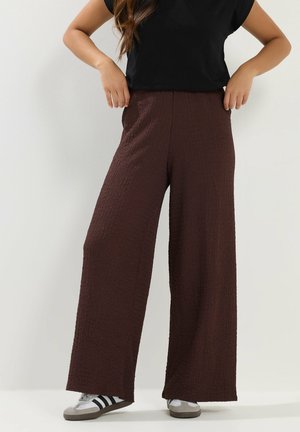 Osoba ubrana w brązowe, szerokie, teksturowane spodnie z wysokim stanem, czarny krótki top i białe sneakersy z czarnymi paskami.