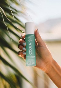 Aqua-grünne Sprühflasche mit einem weißen Deckel, beschriftet mit "COOLA Makeup Setting Sunscreen Spray", enthält SPF 30 und 70 % organische Inhaltsstoffe.