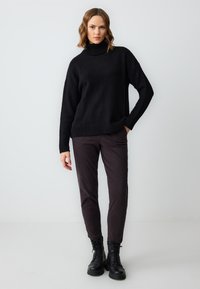Zwarte gebreide coltrui met een losse pasvorm, geribbelde manchetten, gecombineerd met donkerbruine slim-fit broek en zwarte veterlaarzen.