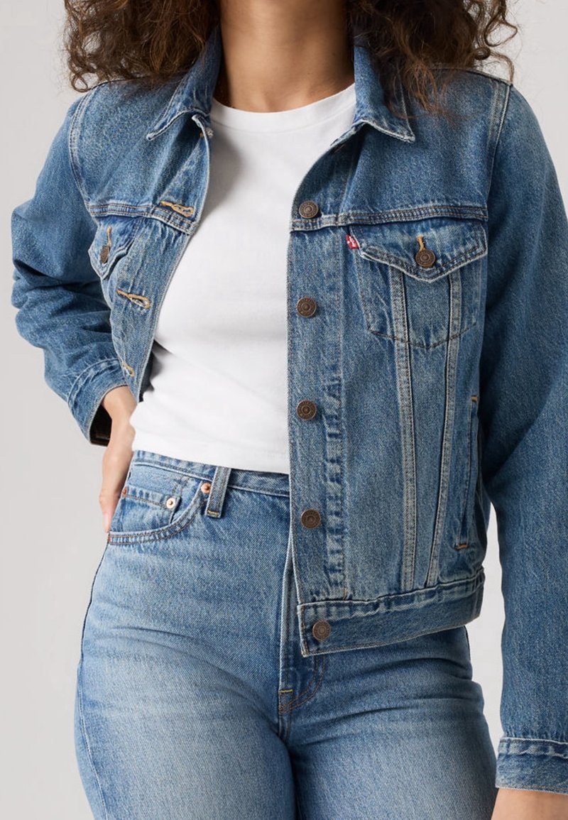 Femme portant une veste en jean bleue sur un t-shirt blanc et un jean bleu, avec une main reposant sur la hanche.