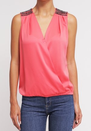 Ärmellose Bluse in Korallenrosa mit V-Ausschnitt und mit Nieten verzierte Schultern, getragen zu blauen Jeans an einer stehenden Person.