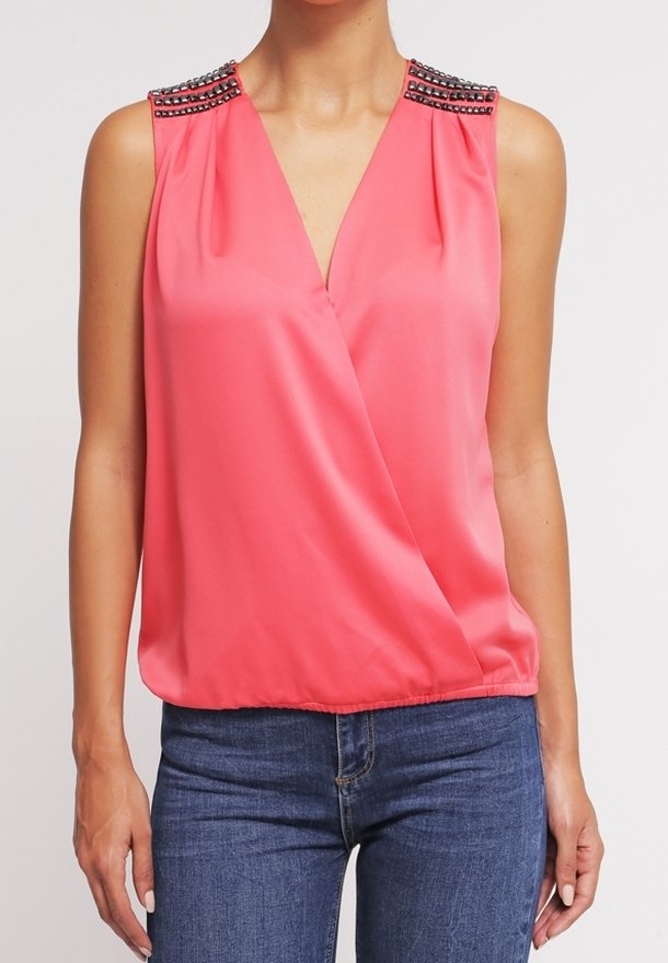 Blouse sans manches rose corail avec col en V et détails cloutés sur les épaules, portée avec un jean bleu sur une personne debout.