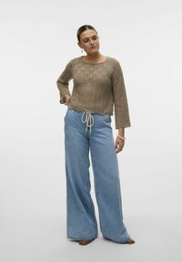 Maglione beige lavorato a maglia con un motivo intricato, abbinato a jeans azzurri a vita alta e gamba ampia, insieme a scarpe marroni. Presenta un cinturino con coulisse.