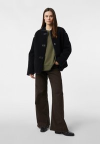 Czarna, teksturowana kurtka z okrągłym kołnierzem i przednimi guzikami, nałożona na oliwkowo zieloną bluzę, zestawiona z brązowymi spodniami o szerokich nogawkach i czarnymi butami.