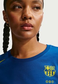 Blå bomulls-T-shirt med rund halsringning och korta ärmar, med en gul FC Barcelona-logga på vänster bröstområde.