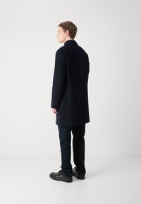 Marineblauer Wollmantel mit tailliertem Schnitt, verlängertem Design, Reverskragen und nahtlosem Rücken. Kombiniert mit dunklen Hosen und schwarzen Schuhen.
