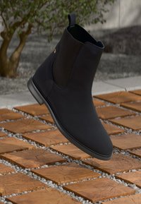 Sort læder ankelstøvle med elastiske sidelapper på trægulv med kvadratiske fliser og små hvidgrus gap, slørede planter i baggrunden.