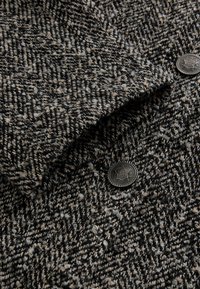 Wełniana tkanina w czarno-beżowy wzór jodełki, o teksturowanej powierzchni oraz ozdobiony metalowy guzik z misternym zdobieniem.