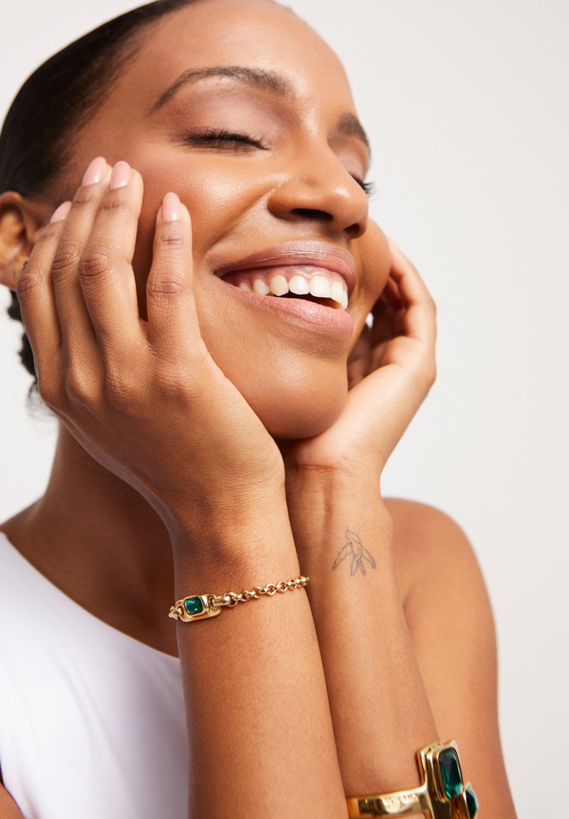 Bracelet en or avec un détail de pierre précieuse verte sur un poignet. Le teint de peau est chaud. Présente un tatouage discret sur la partie intérieure du bras. Fond neutre.