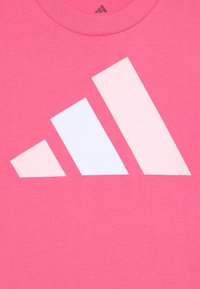 Rozā kokvilnas t-krekls ar lielu, minimālisma stila baltu logo, kas sastāv no trim leņķainām formām dažādos izmēros un orientācijās.