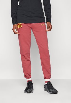 Person iført røde joggers med gult Nike-logo, sort langærmet trøje, sorte sportssko og rød-hvide strømper, står indendørs.