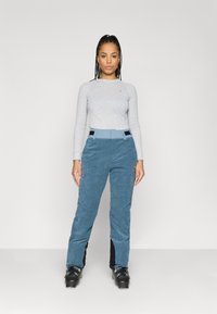 Top de mangas longas cinza claro combinado com calças de veludo côtelê turquesa com acabamento texturizado, detalhes laterais pretos e botas de esqui.