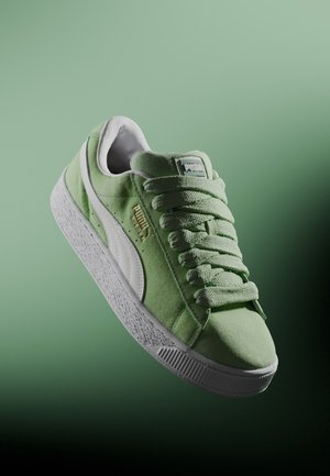 Baskets Puma en daim vert clair avec une bande blanche et une semelle blanche épaisse texturée, présentées en angle sur un fond dégradé vert.