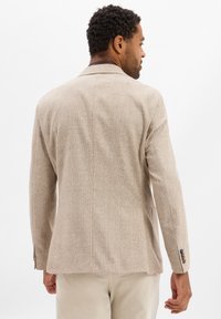 Mann in beigem kariertem Blazer und hellen Hosen, von hinten betrachtet vor einem weißen Hintergrund.