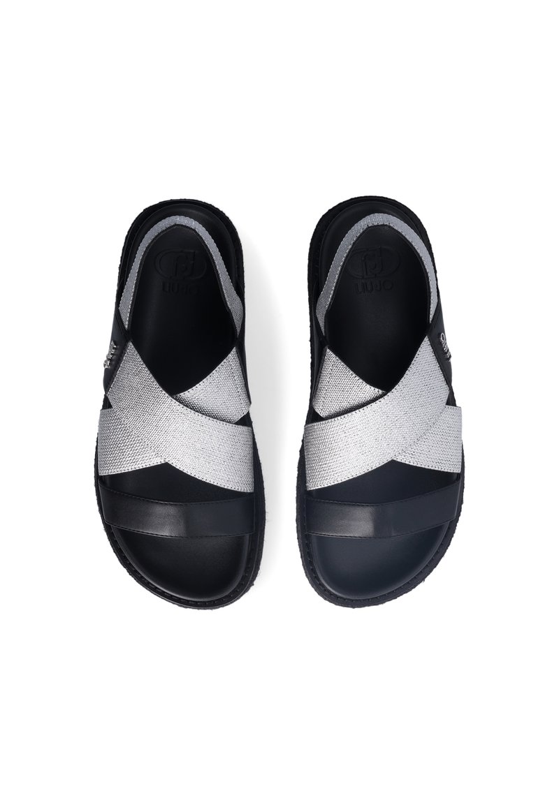 Sandales glissantes noires et argentées avec des bandes croisées, présentant une texture tissée et des accents en cuir lisse. Semelles plates en caoutchouc pour plus de stabilité.