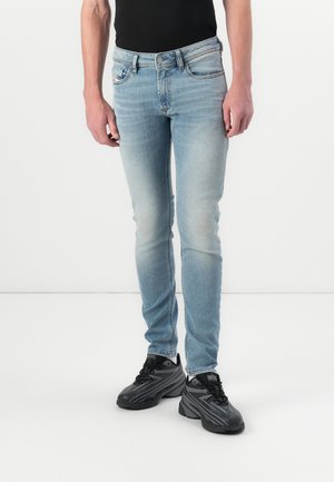 Homme portant un jean slim bleu clair et des baskets noires, debout devant un fond blanc, les mains détendues le long du corps.